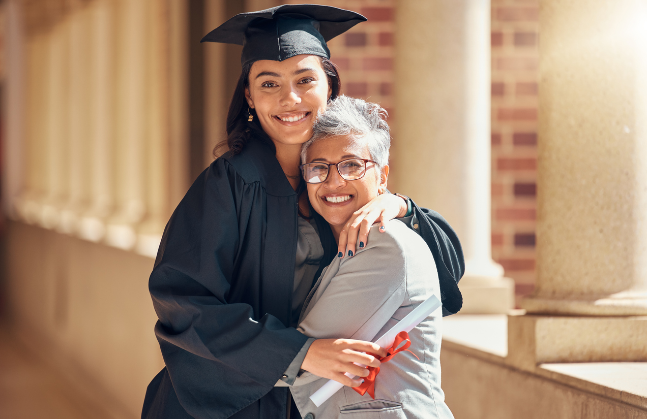 Graduate in cap and gown hugging an older woman while holding a diploma.