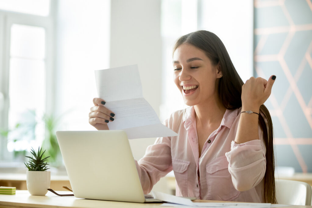 Student smiling while reading a letter at a laptop in a bright room.