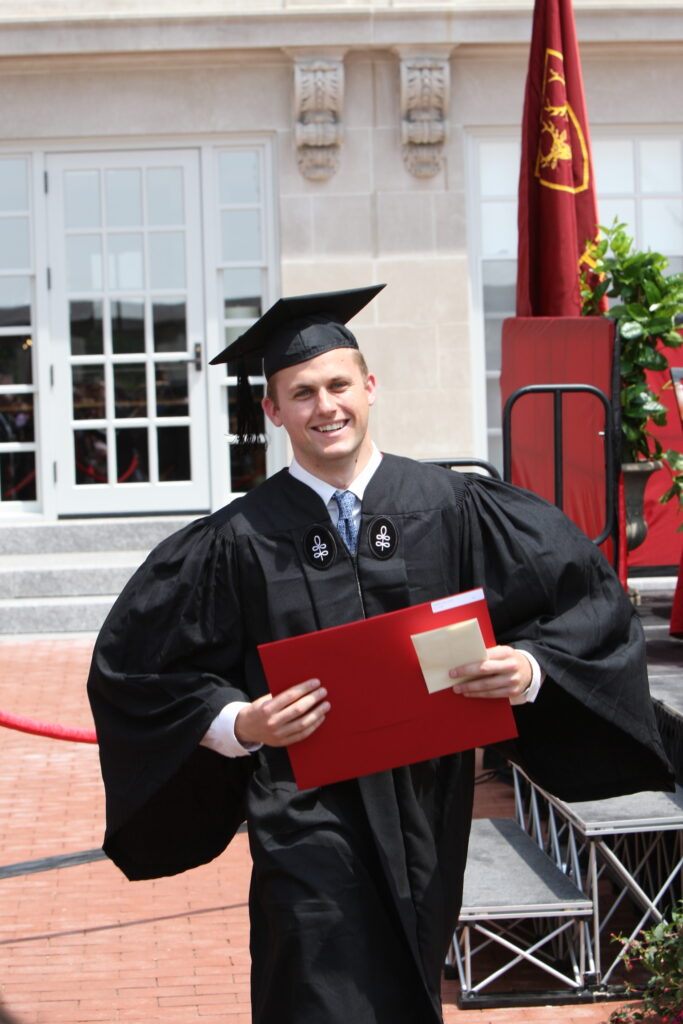 Graduate in cap and gown holding a diploma outside a campus building.