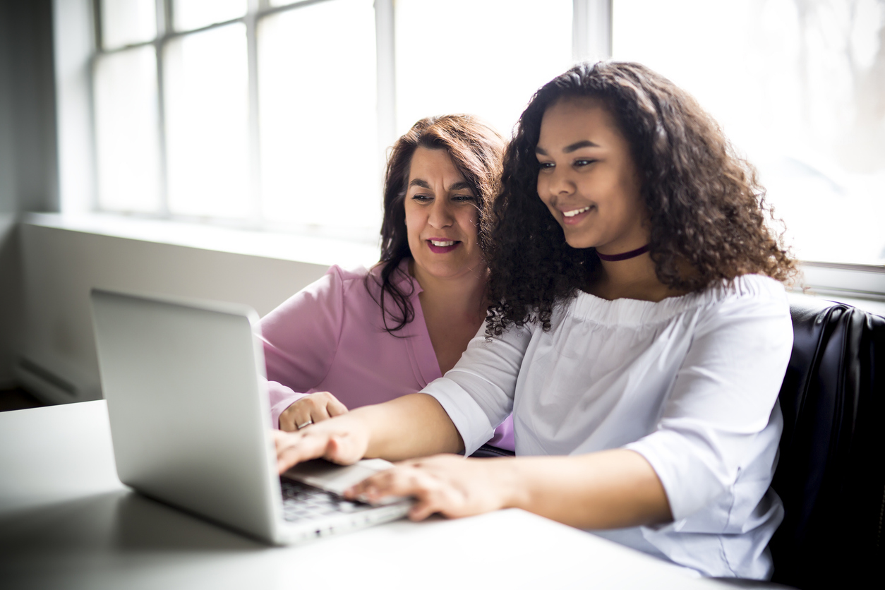 Two women working together on a laptop at a table by a window.