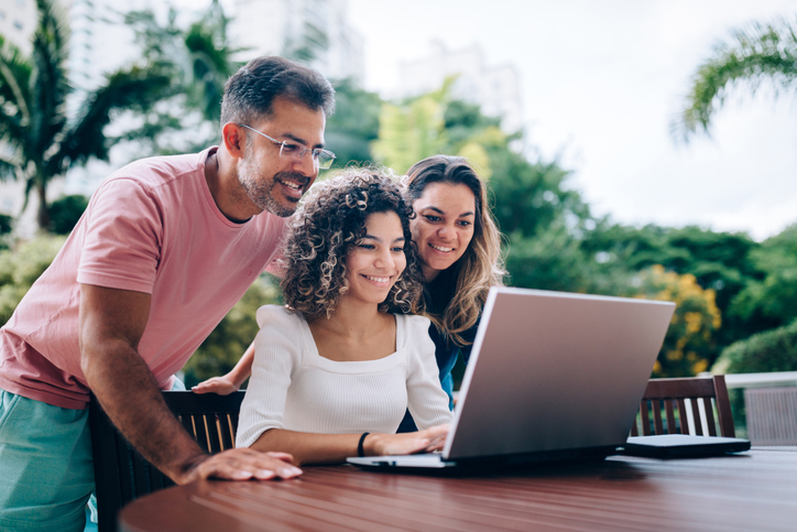 Parents watching their child while using her laptop at an outdoor table.