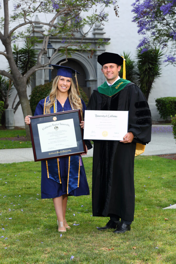 Two graduates in caps and gowns holding diplomas on a lawn in front of a campus building.