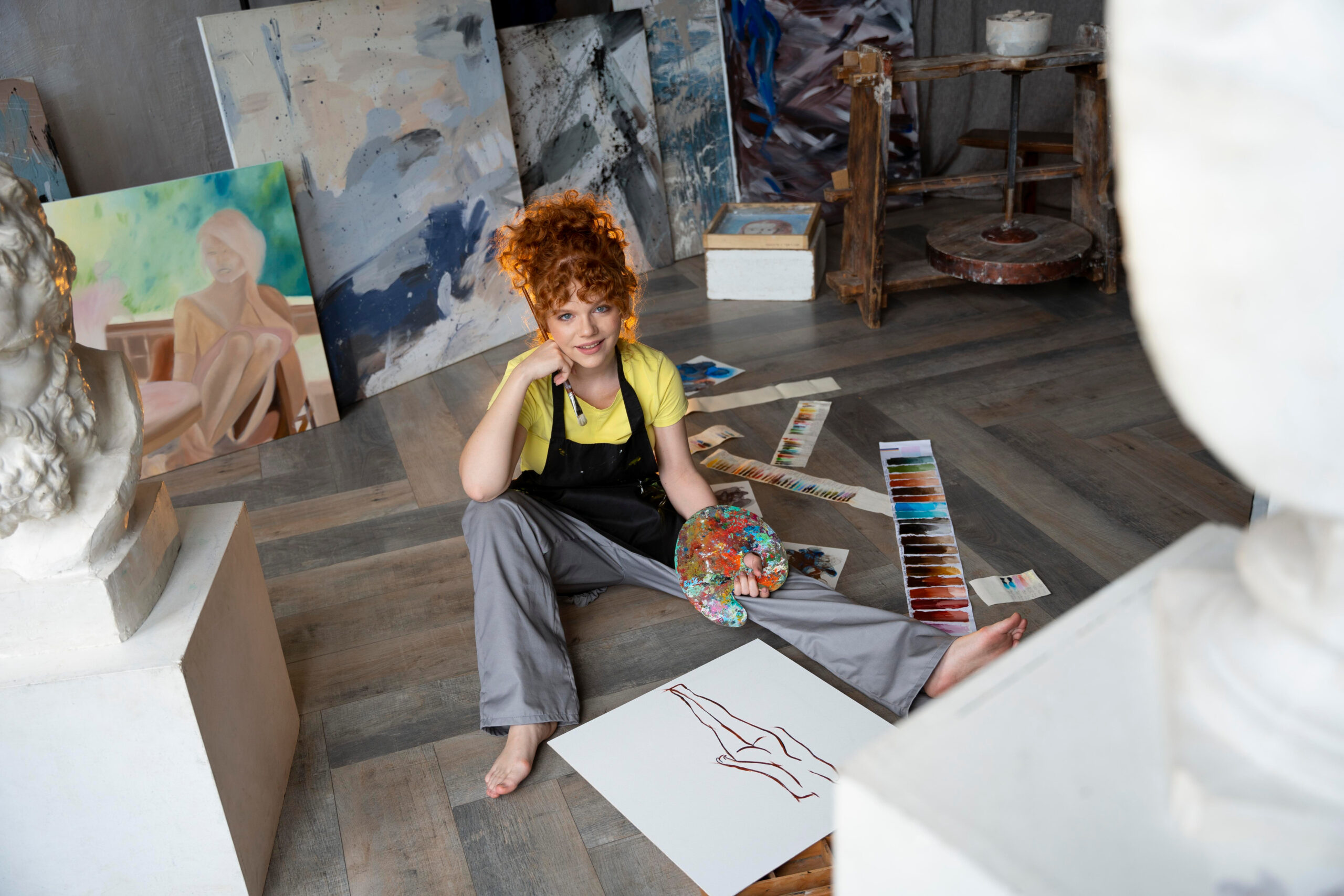 A woman with red curly hair is sitting on the floor while painting in an art room full of paintings and sculptures.