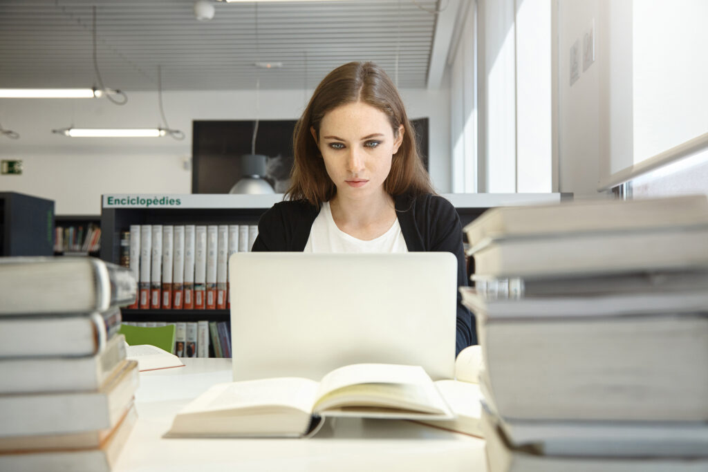 A student studying from her laptop at a desk surrounded by stacked books.