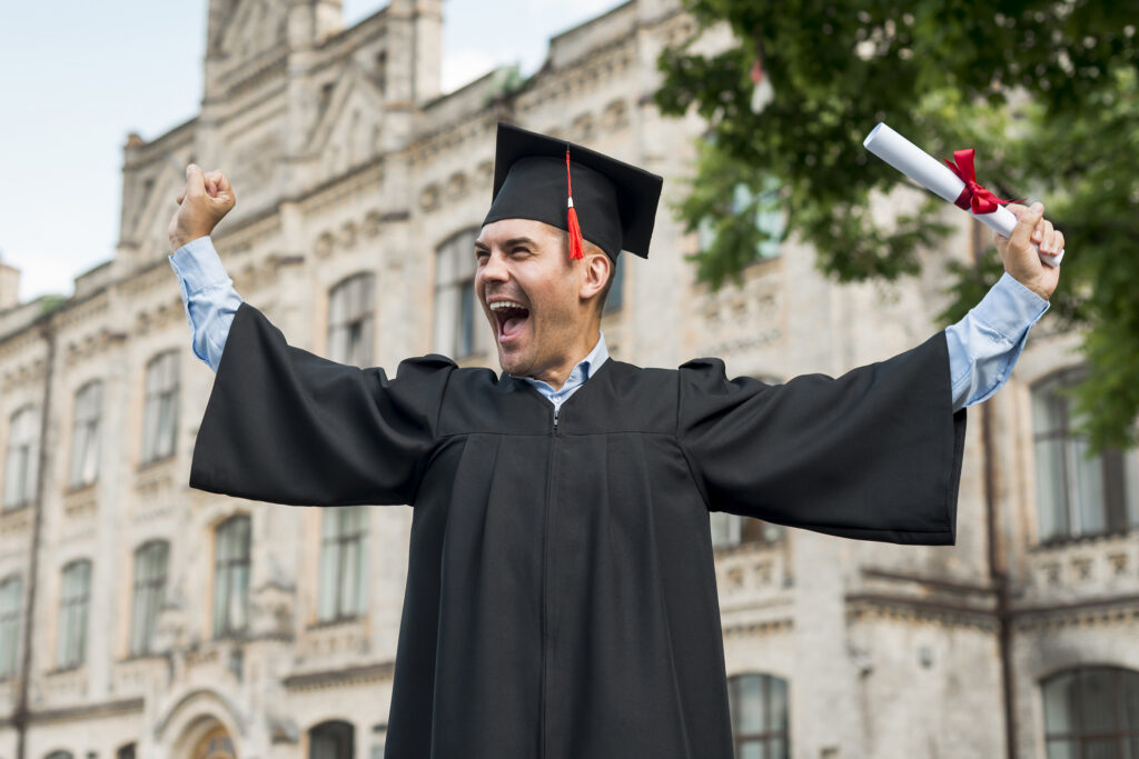 Graduate in cap and gown cheering while holding a diploma outdoors.