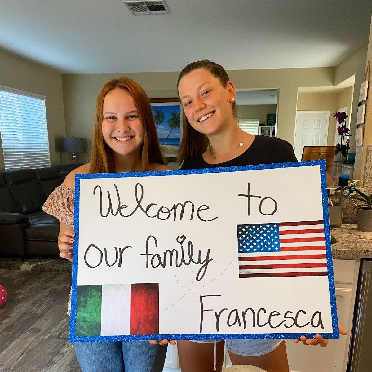 Two women holding a welcome sign with U.S. and Italian flags in a home.