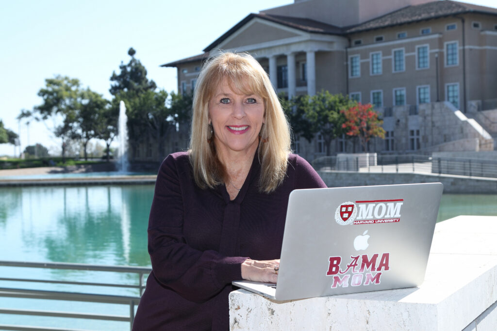 Shellee is wearing a deep purple shirt while facing her laptop in the outdoors with a lake and a fountain in the background.