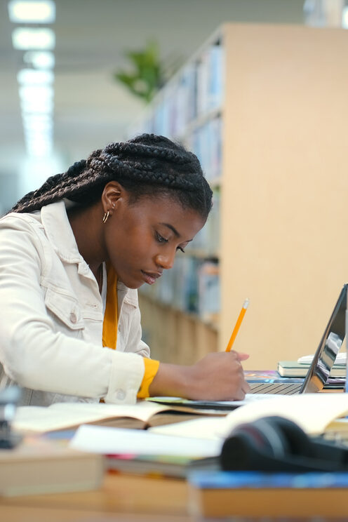 Student writing notes at a desk while studying in a library.