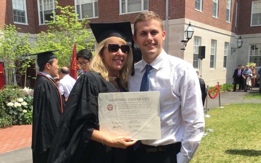 Graduate holding a diploma while standing with an adult outside a campus building.