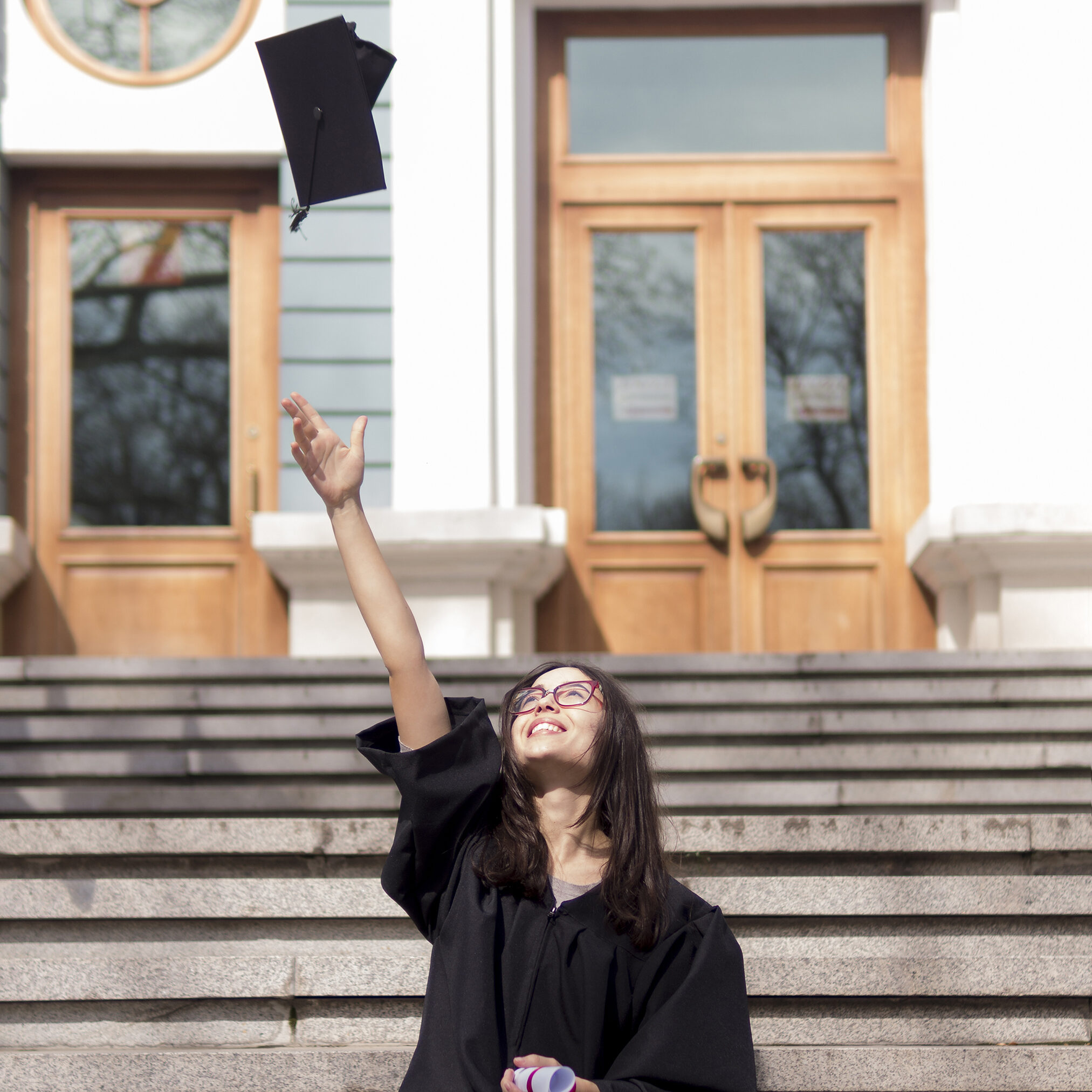 Graduate in cap and gown tossing a hat into the air while sitting on outdoor steps.