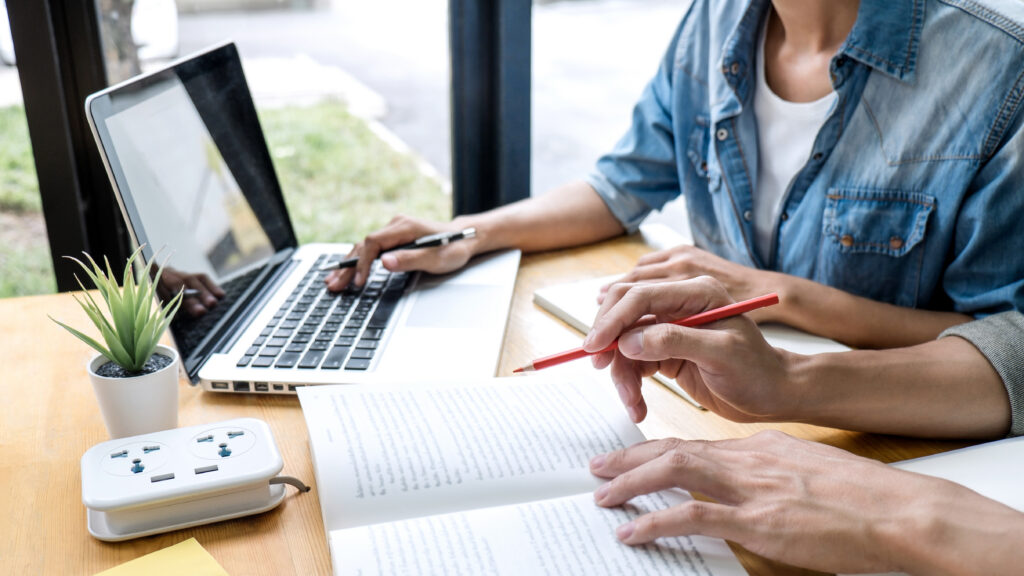 Two people working together at a desk with a laptop and paperwork, pointing at a document while taking notes.