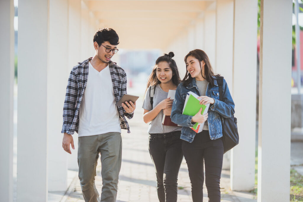 Three college students walking together on campus while looking at a smartphone and carrying books.