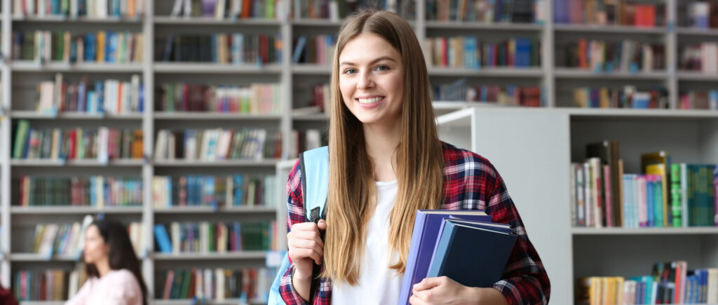 Student standing in a library holding books and smiling with shelves of books in the background.