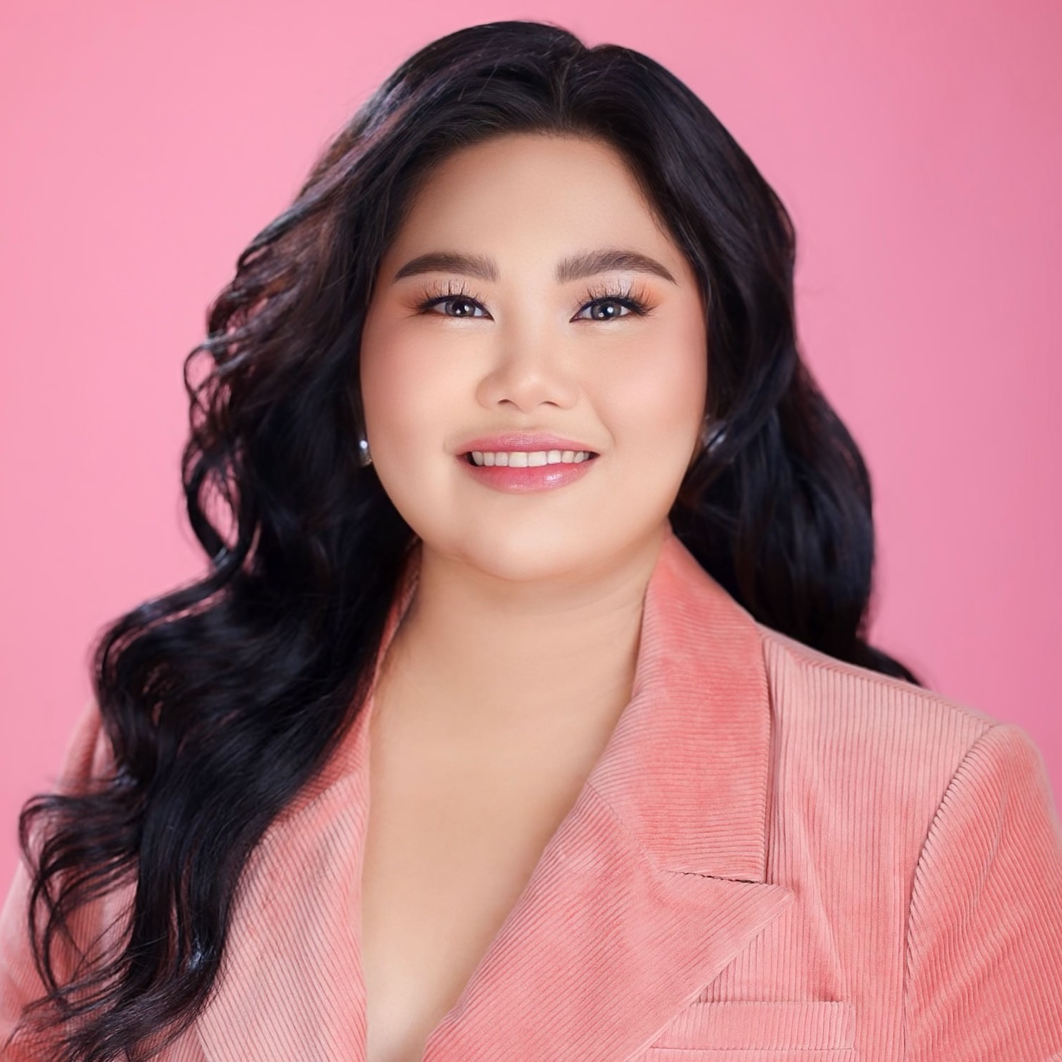 Professional headshot of a smiling woman with long dark hair wearing a pink blazer against a soft pink background.