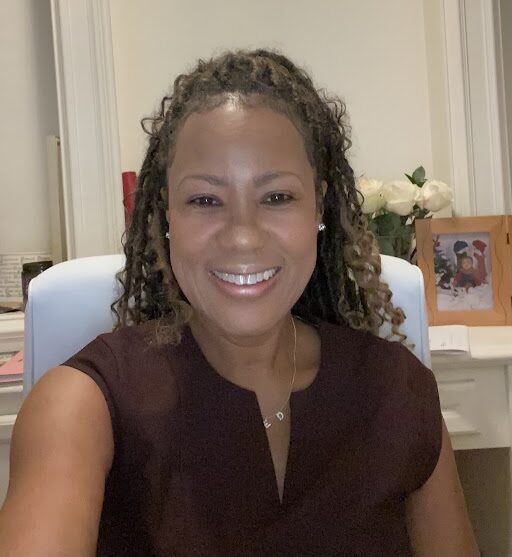 Smiling woman with curly hair seated at a desk in a home office, with a framed photo and flowers in the background.