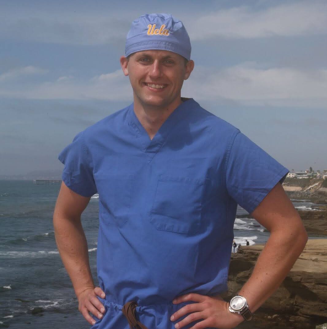 Man wearing blue medical scrubs and a UCLA cap standing with hands on hips by the ocean shoreline.