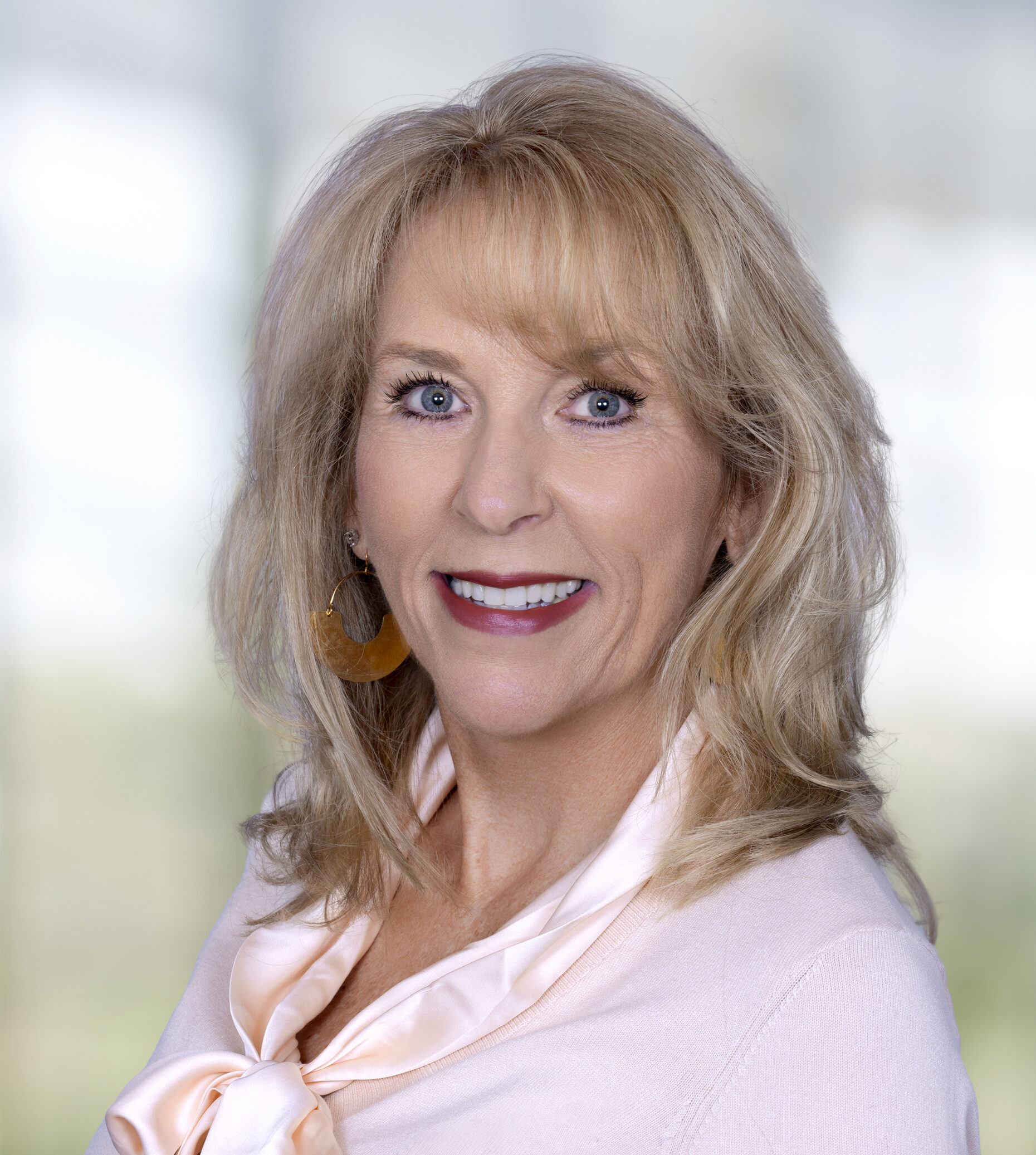 Professional headshot of a smiling blonde woman wearing a light pink blouse and gold earrings against a softly blurred background.