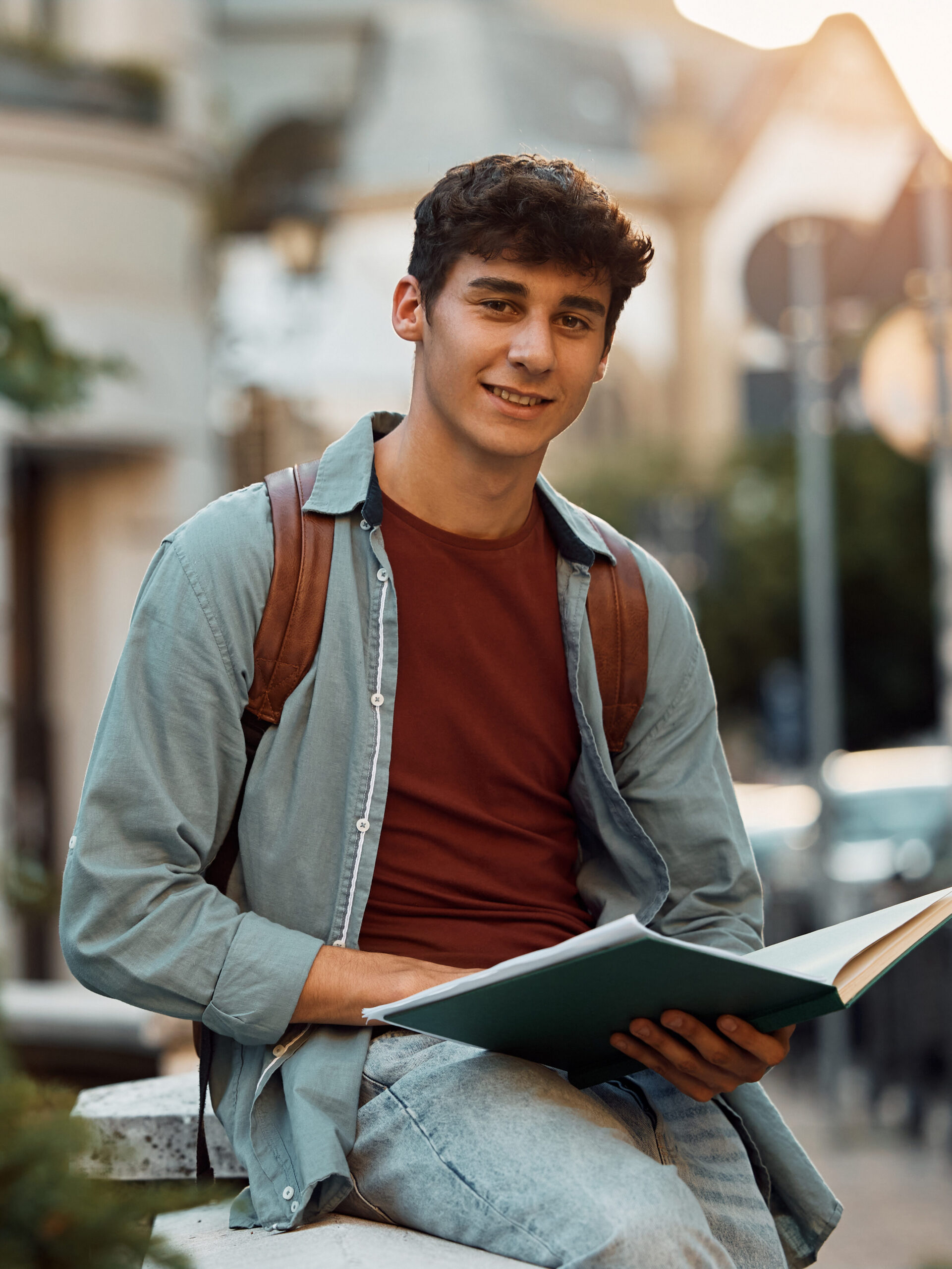 Smiling college student reading book at campus and looking at camera.