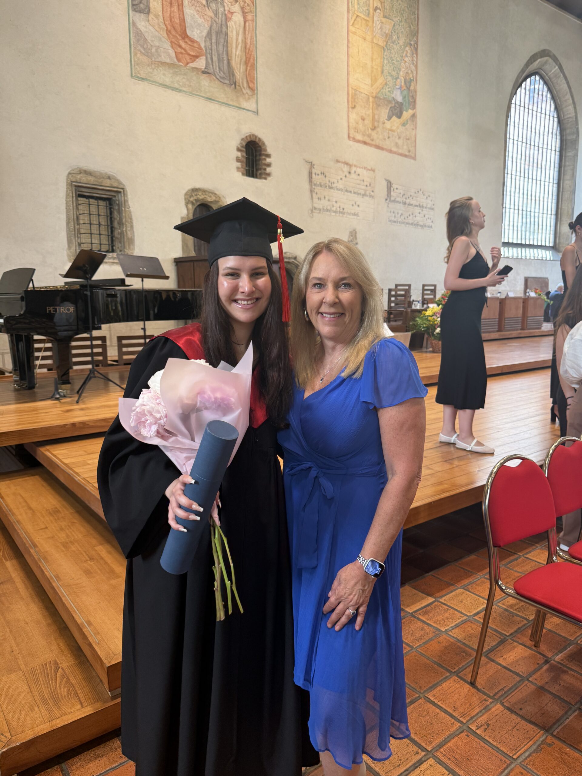 Graduate in cap and gown holding bouquet standing with woman in blue dress at indoor graduation ceremony.