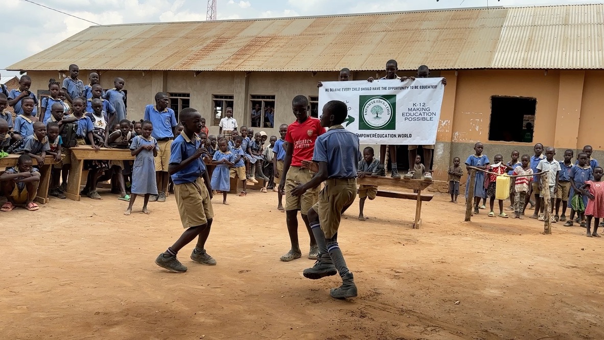 Students in school uniforms performing a game or dance in a rural schoolyard while other children watch near a classroom building.