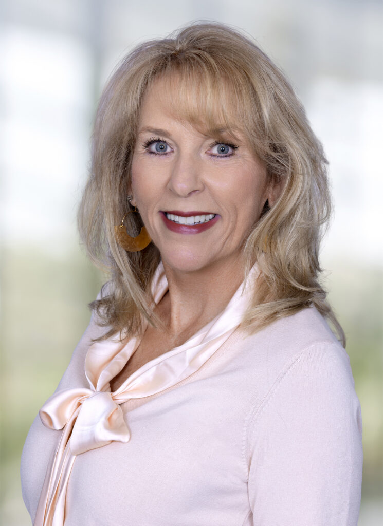 Professional headshot of a smiling blonde woman wearing a light pink blouse and gold earrings against a softly blurred background.