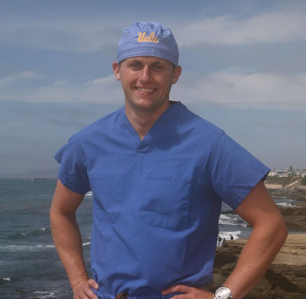 Man wearing blue medical scrubs and a UCLA cap standing with hands on hips by the ocean shoreline.