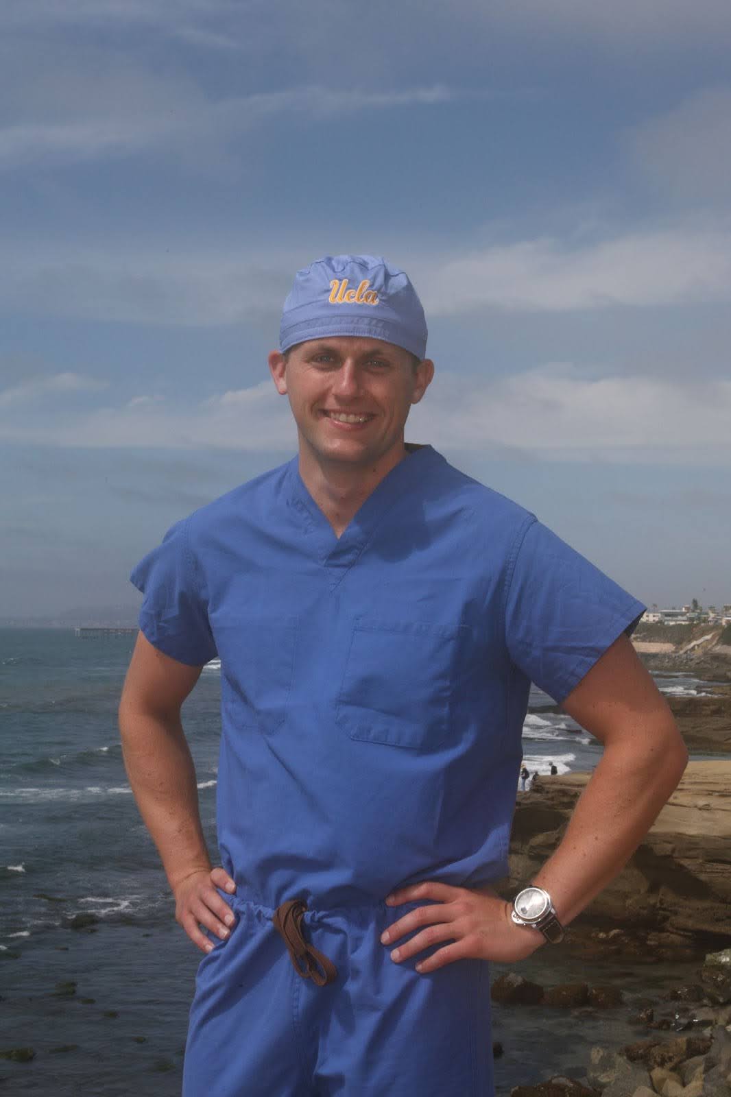 Man wearing blue medical scrubs and a UCLA cap standing with hands on hips by the ocean shoreline.