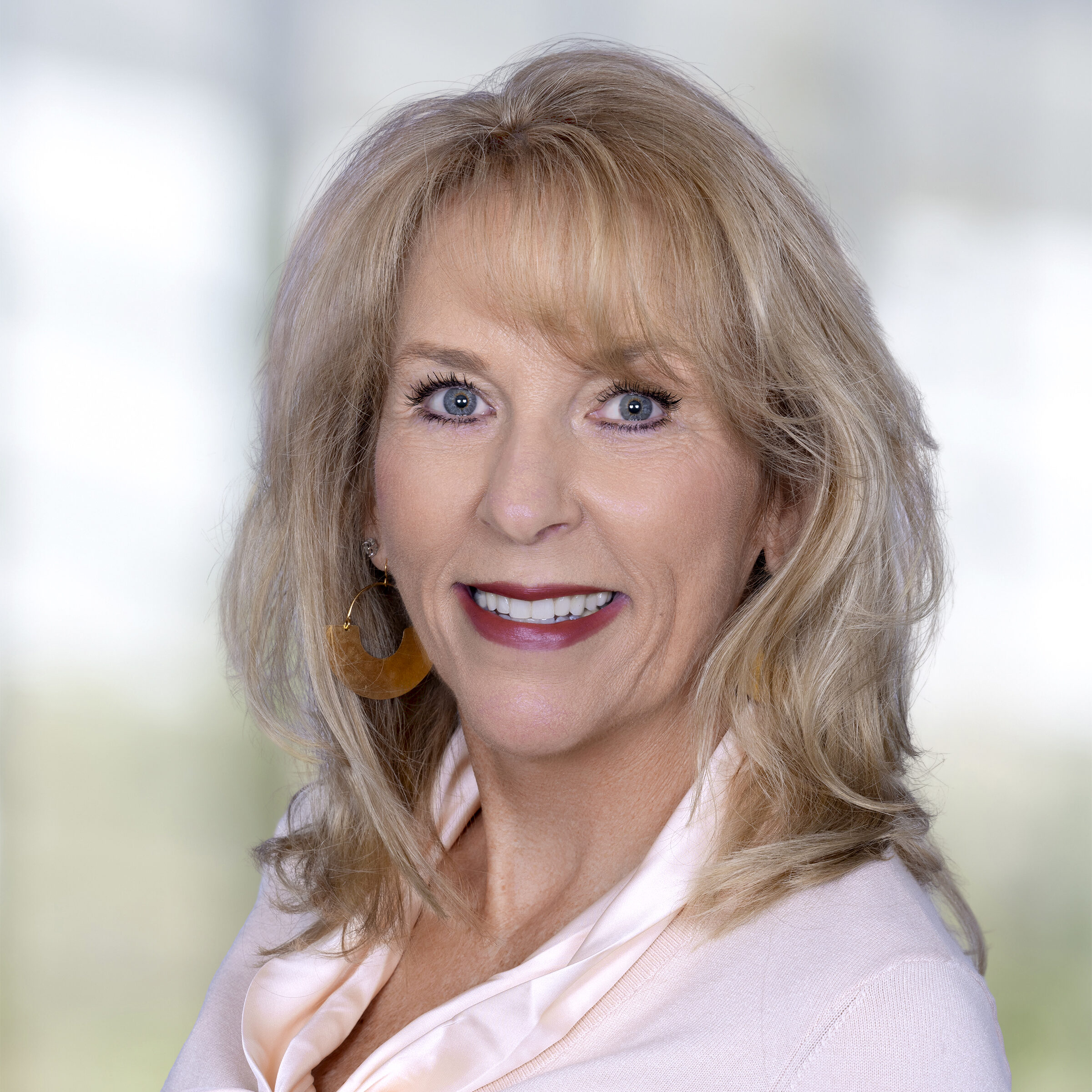 Professional headshot of a smiling blonde woman wearing a light pink blouse and gold earrings against a softly blurred background.