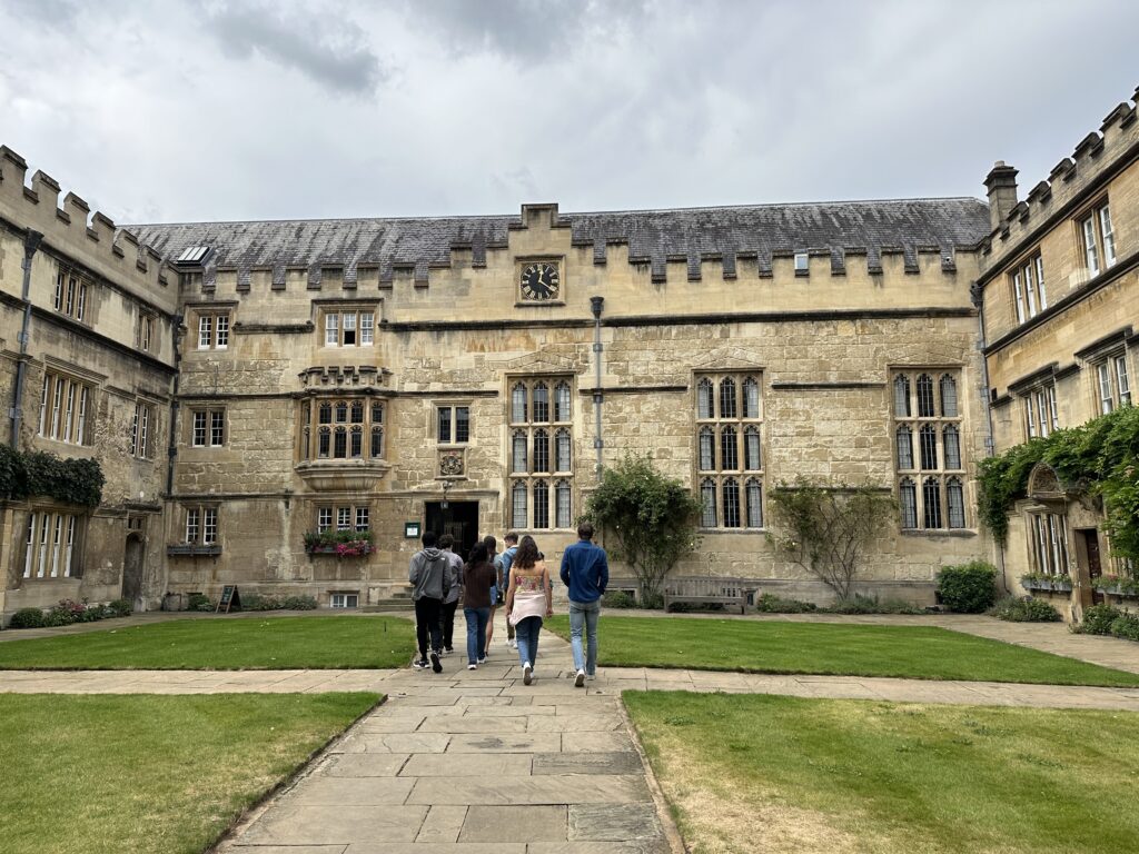 A group of people walks along a stone pathway toward a historic academic building constructed from light-colored stone.