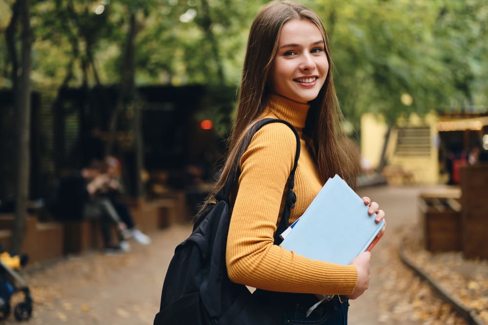 Student carrying notebooks and a backpack while walking outdoors on campus.