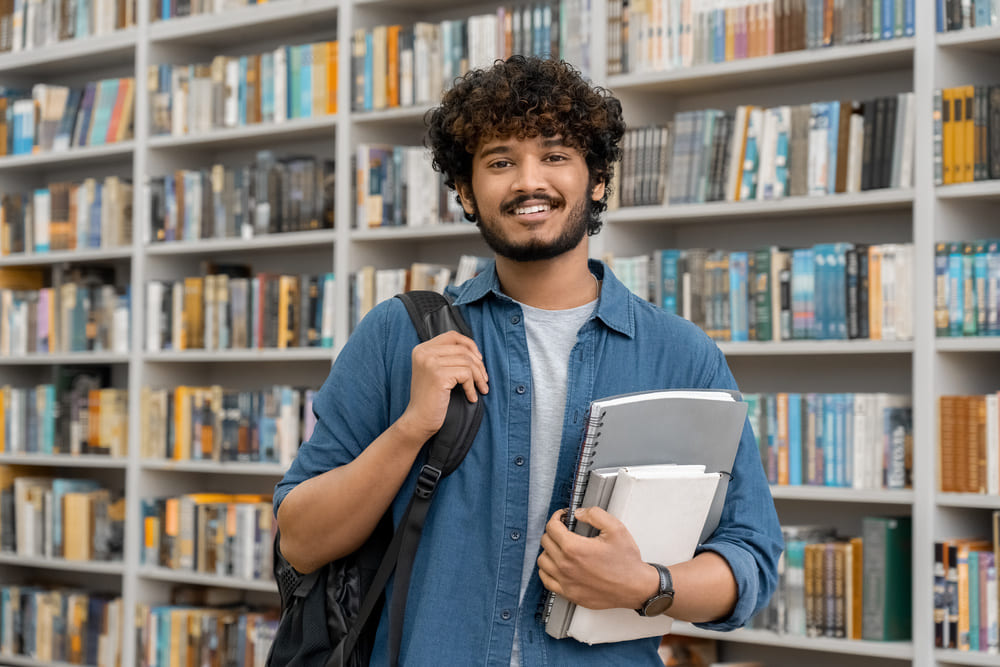 Student with a backpack holding books while standing in a library surrounded by shelves.