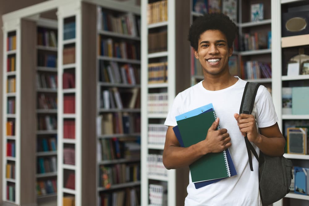 Student holding books and smiling while standing in front of bookshelves in a library.